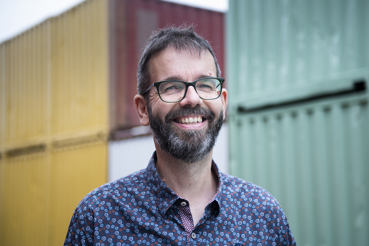 Lachende man met baard en bril, in een overhemd met bloemenprint, voor gekleurde zeecontainers